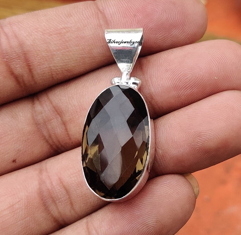 K&ouml;nnte beinhalten: Ein silberner Anh&auml;nger mit einem gro&szlig;en, facettierten, ovalen Rauchquarz-Edelstein. Der Anh&auml;nger hat eine &Ouml;se mit dem eingravierten Text "Silverjewelry20" auf der R&uuml;ckseite.