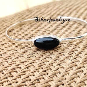 May include: A silver bangle bracelet featuring a black oval gemstone. The bracelet is displayed on a woven brown surface.