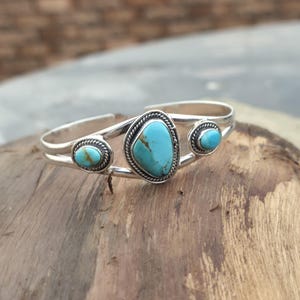 May include: A silver cuff bracelet with three turquoise stones. The central stone is oval-shaped, and the two flanking stones are round. The bracelet has a braided rope design around the stones.