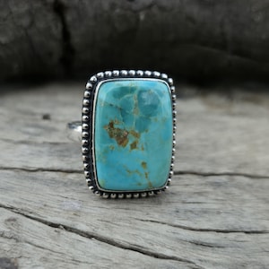 Peut inclure: Bague en argent ornée d'une grande pierre turquoise rectangulaire. La turquoise présente un motif moucheté de bleu et de brun. Le sertissage de la bague est en argent avec une bordure perlée, pour un look vintage ou artisanal.