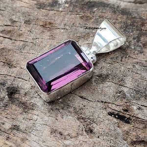 Puede incluir: Un colgante de plata con una gran piedra preciosa rectangular de color púrpura. El colgante está engastado en un bisel de plata y tiene un aro para sujetarlo a una cadena.