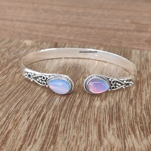May include: Silver bangle bracelet with two teardrop-shaped opal stones. The stones are a mix of pink, blue, and white.