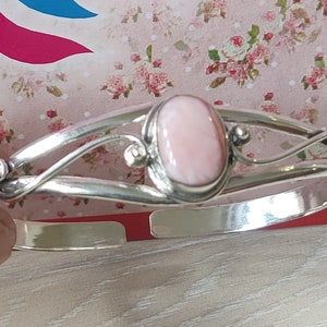 Op de afbeelding: Een zilverkleurige manchetarmband met een grote, ovale, lichtroze edelsteen in het midden. De armband heeft een decoratief, vloeiend ontwerp met kleine, ronde accenten en een open, verstelbaar ontwerp. De steen is ongeveer 2 cm lang.