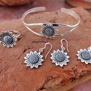 May include: A silver sunflower jewelry set with a bangle bracelet, ring, earrings, and pendant. The sunflower design is detailed and intricate.