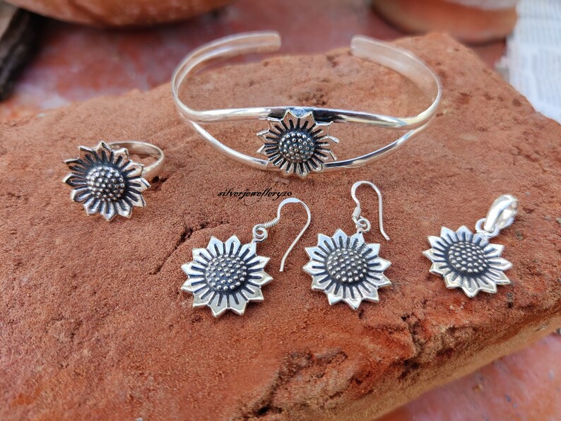 May include: A silver sunflower jewelry set with a bangle bracelet, ring, earrings, and pendant. The sunflower design is detailed and intricate.