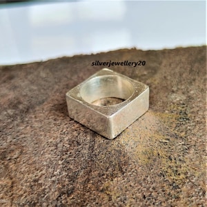 Pode incluir: Um anel quadrado de prata com um acabamento liso e polido. O anel é grosso e tem uma banda larga.