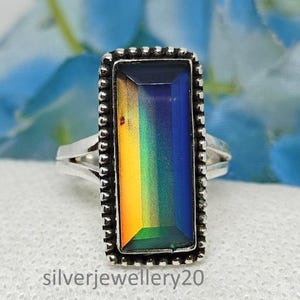May include: A silver ring with a rectangular, color-changing stone. The stone transitions from yellow to green to blue. The ring has a beaded border and a split shank. The text "silverjewellery20" is at the bottom.