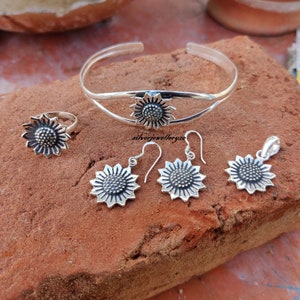 May include: A silver sunflower jewelry set with a bangle, ring, earrings, and pendant. The jewelry is made of sterling silver and features a detailed sunflower design.