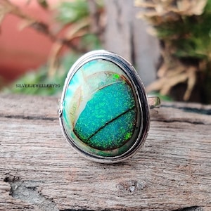 May include: Oval-shaped silver ring with a large, iridescent gemstone. The gemstone features shades of teal, green, and hints of gold. The ring is set on a wooden surface. The text "SILVERJEWELLERY20" is visible on the ring.