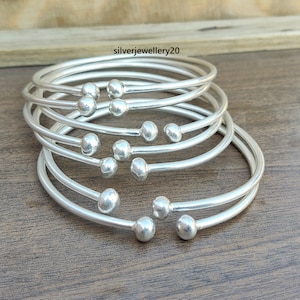May include: A set of eight silver bangles with round ball accents. The bangles are stacked on top of each other on a wooden surface.