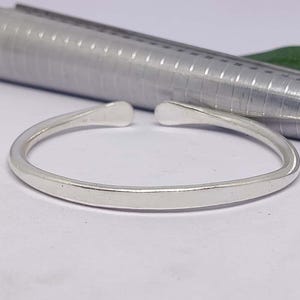 Puede incluir: Un brazalete abierto de color plateado con extremos redondeados. El brazalete está enfocado, con un medidor de anillos plateado y una hoja verde en el fondo. El brazalete está sobre una superficie blanca.