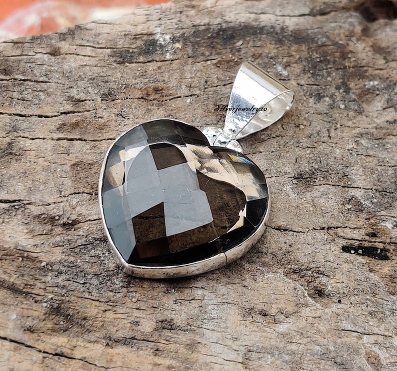 Pode incluir: Um pingente de prata em forma de cora&ccedil;&atilde;o com uma pedra preciosa de quartzo fumado escuro. O pingente est&aacute; sobre uma superf&iacute;cie de madeira.