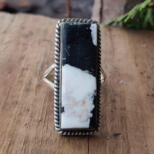 Könnte beinhalten: Rechteckiger Ring mit einem schwarz-weißen Stein und einer silberfarbenen Fassung. Der Stein hat ein marmoriertes Aussehen, mit einer Mischung aus schwarzen und weißen Abschnitten. Die Fassung des Rings hat ein gedrehtes Seildesign.