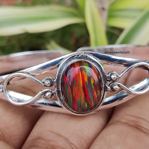 May include: A silver cuff bracelet with an oval, multi-colored gemstone at the center. The gemstone displays streaks of red, orange, yellow, and green. The bracelet has an open design with decorative swirls and small silver beads.