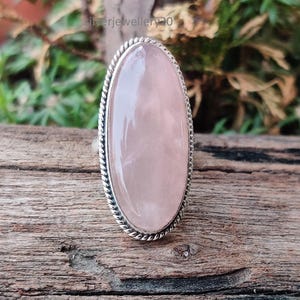 Puede incluir: Un anillo con una gran piedra ovalada de cuarzo rosa. La piedra rosa claro está engastada en una banda de metal plateado con un diseño de cuerda trenzada alrededor del borde. El anillo se exhibe sobre una superficie de madera con un fondo verde borroso.