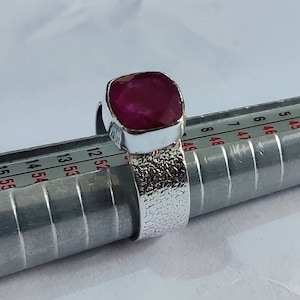 Może przedstawiać: Srebrny pierścionek z fakturowanym obrączką i kwadratowym, ciemnoróżowym rubinem. Pierścionek jest rozmiarowany na metalowym mierniku pierścionków.