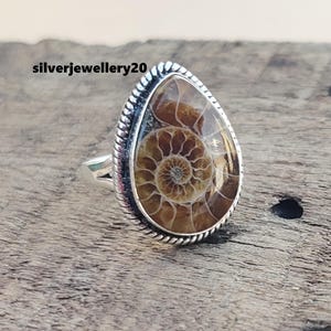 May include: A silver ring featuring a teardrop-shaped ammonite gemstone. The ammonite is brown and has a spiral pattern.