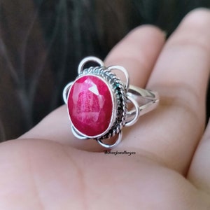 May include: A silver ring with a red ruby gemstone set in a floral design. The ring is on a hand.