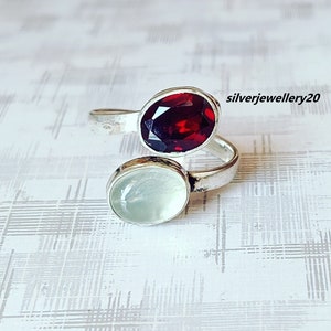 May include: A silver ring featuring two oval-shaped gemstones. The larger stone is a deep red garnet, and the smaller stone is a pale green moonstone.