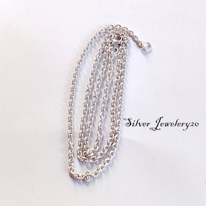Può includere: Una collana a catena d'argento con una chiusura a moschettone. La catena è composta da piccoli anelli rotondi. La catena è piegata a metà e appoggiata su una superficie bianca. Il testo "Silver Jewellery20" è visibile nell'angolo in basso a destra dell'immagine.