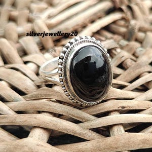 Può includere: Un anello d'argento con una pietra ovale nera incastonata in una montatura a perline. L'anello è su uno sfondo intrecciato.