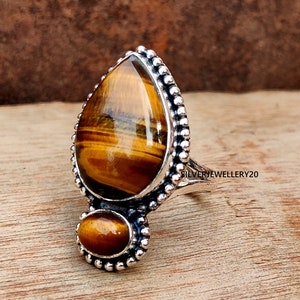 May include: A silver ring featuring two teardrop-shaped tiger's eye stones. The larger stone is set in a bezel setting with a beaded border. The smaller stone is set in a similar setting below the larger stone. The ring band is plain and silver.