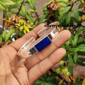 May include: A silver cuff bracelet with a rectangular blue lapis lazuli stone set into the band. The bracelet is open and is being held in a hand.