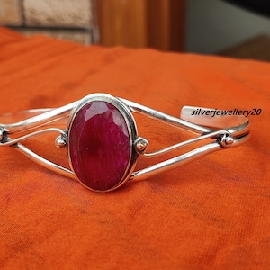 Könnte beinhalten: Ein silbernes Armreif-Armband mit einem großen, ovalen, tief roten Edelstein in der Mitte. Das Armband hat ein zartes, gedrehtes Design und ist mit kleinen silbernen Perlen verziert.