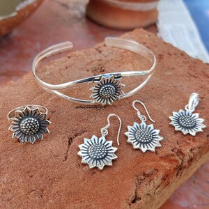 May include: A silver sunflower jewelry set with a bangle bracelet, ring, earrings, and a pendant. The jewelry is made of sterling silver and features a detailed sunflower design.