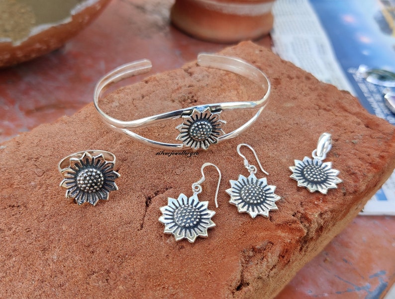 May include: A silver sunflower jewelry set with a bangle bracelet, ring, earrings, and a pendant. The jewelry is made of sterling silver and features a detailed sunflower design.