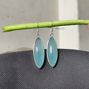 May include: A pair of silver dangle earrings with light blue oval-shaped chalcedony gemstones. The earrings are hanging from a green stem.