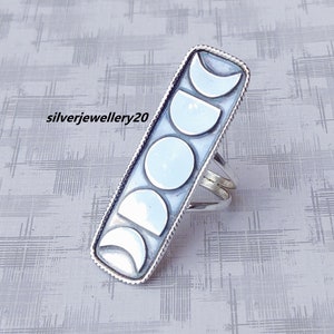 Puede incluir: Un anillo de plata con un diseño rectangular que muestra las fases lunares. El anillo tiene un borde texturizado y el texto "silverjewellery20" es visible. Las fases lunares se representan en diferentes formas, desde la media luna hasta la luna llena.