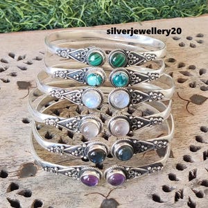 May include: A set of six silver bangles with different colored gemstones. The bangles are stacked on top of each other and feature intricate designs. The gemstones include green, blue, white, pink, and purple.