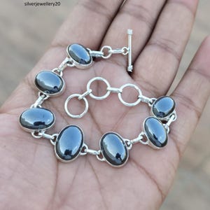 Puede incluir: Una pulsera de plata con piedras preciosas ovaladas de color gris oscuro. Las piedras están engastadas en eslabones individuales de plata, conectados por pequeños anillos de plata. La pulsera tiene un cierre de palanca. La joya se muestra en una mano.