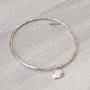 Könnte beinhalten: Ein silbernes Armreif-Armband mit gehämmerter Oberfläche und einem kleinen Herz-Anhänger. Das Armband liegt auf einem hellen Holzuntergrund.