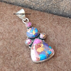 May include: A silver pendant with a triangular turquoise and pink stone surrounded by smaller round stones. The pendant is on a brown surface.