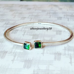 May include: A silver bangle bracelet with two square green gemstones. The bracelet is open and has a silver inscription that reads "925 silverjewellery20".