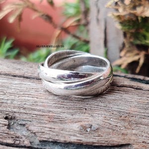 Könnte beinhalten: Ein silberfarbener Ring mit drei ineinandergreifenden Bändern. Der polierte Ring reflektiert das Licht und erzeugt ein glänzendes Aussehen. Der Ring liegt auf einer rustikalen Holzoberfläche. Der Text "SILVERJEWELLERY20" ist sichtbar.