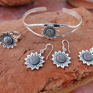 May include: A silver sunflower jewelry set with a bangle bracelet, ring, earrings, and pendant. The sunflower design is detailed and intricate.