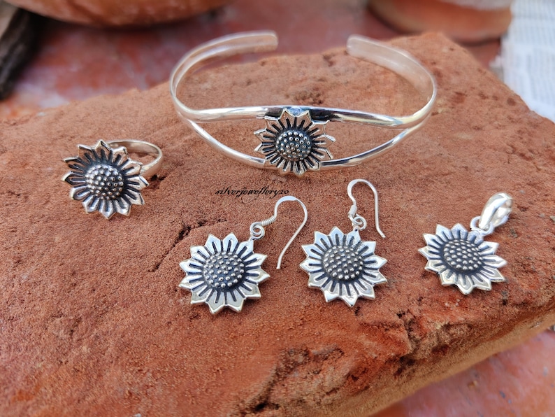 May include: A silver sunflower jewelry set with a bangle bracelet, ring, earrings, and pendant. The sunflower design is detailed and intricate.