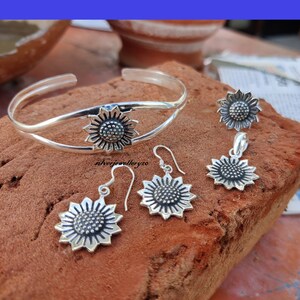 May include: A silver sunflower bracelet, a pair of silver sunflower earrings, and a silver sunflower pendant necklace. The jewelry is on a brown textured surface.