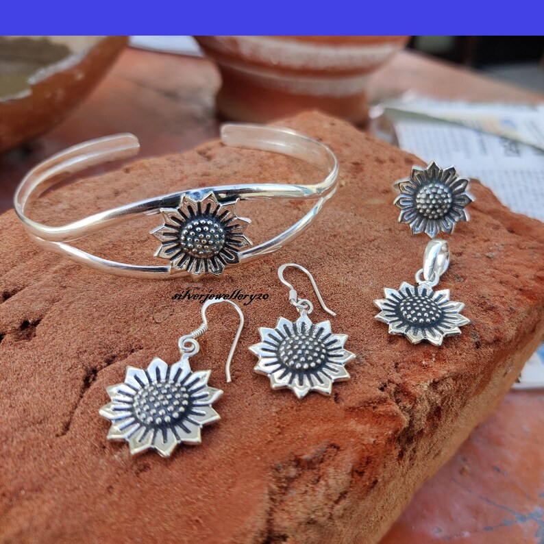 May include: A silver sunflower bracelet, a pair of silver sunflower earrings, and a silver sunflower pendant necklace. The jewelry is on a brown textured surface.
