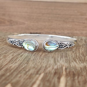 May include: A silver bangle bracelet with two iridescent abalone shell accents. The bracelet is open and has a delicate design.