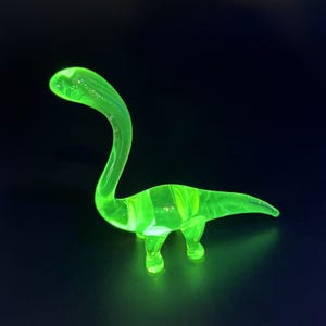 May include: A neon green glass dinosaur figurine with a long neck and a curved tail. The figurine is transparent and glows brightly against a dark background. The dinosaur is a Brontosaurus.