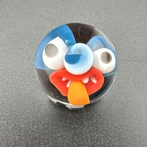 May include: A round, glass art piece with a cartoon face design. The face features blue, white, red, and orange colors. The eyes are white with gray pupils, and the mouth is red with an orange tongue.