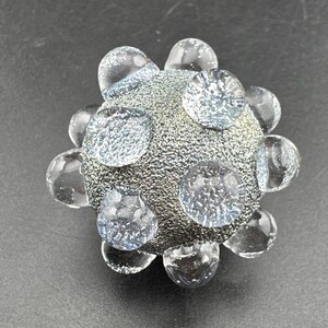 May include: A spherical glass object with a textured, metallic silver finish. The surface is adorned with multiple clear, raised glass orbs that protrude from the center, creating a three-dimensional, studded appearance with a reflective quality.