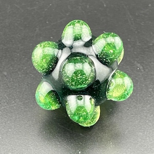 Pode incluir: Uma conta de vidro esférica com uma base escura e translúcida, apresentando seis protuberâncias arredondadas. A superfície do vidro é infundida com partículas finas de glitter verde cintilante por toda a estrutura, criando um efeito reflexivo.