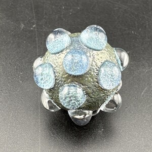 May include: A spherical glass bead with a textured, matte olive-green surface. The bead is adorned with several raised, smooth, translucent light blue circular glass cabochons that have a sparkling, glittery appearance embedded within them.