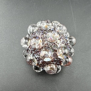 May include: A circular glass bead featuring a cluster of clear, spherical glass bubbles arranged over a dark, textured base. The surface displays iridescent flecks and internal patterns within each bubble, creating a multi-dimensional appearance.