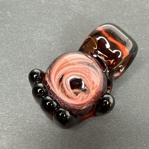 May include: A dark red glass pendant with a swirl of pink and white glass inside. The pendant has a loop at the top for hanging and five black glass beads around the bottom.